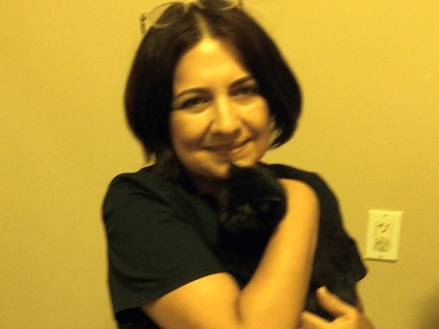 A woman with short hair wearing a black shirt smiles while holding a small black cat close to her chest in a room with beige walls. The image highlights the joy of adopting pets.