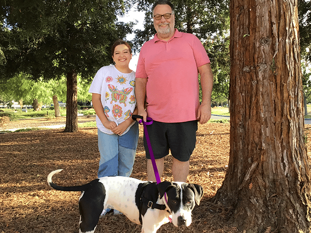 A man in a pink polo shirt and glasses stands next to a young woman in a floral shirt, both smiling. They are in a park with trees in the background, and a black and white dog on a leash is sitting beside them, symbolizing successful adopted pets.
