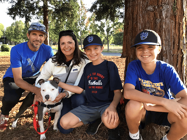 A family of four, including two boys, poses with an adopted dog in a park. The woman is holding the dog on a leash, and they are all smiling. The background features tall trees and a sunny day, highlighting the joyful moment of pet adoption.