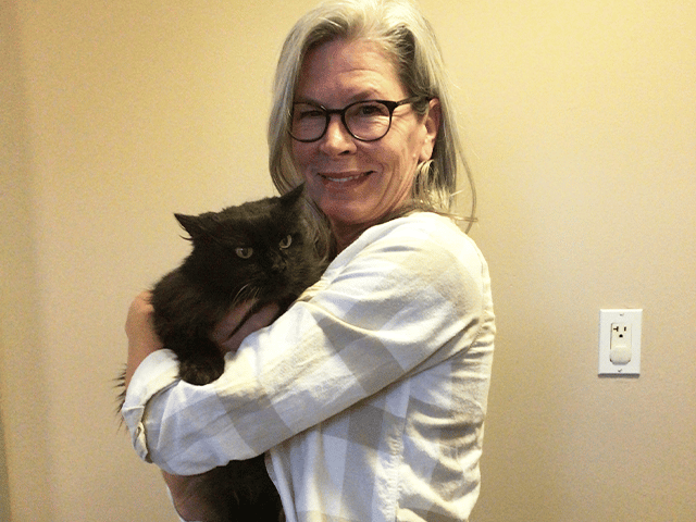 A woman with long gray hair and glasses holds a black cat close to her chest, smiling gently. The background is a light beige color, suggesting a home environment, highlighting the theme of adopted pets.