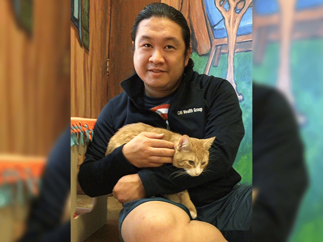 A man seated on the floor in a room with colorful walls holds an orange tabby cat on his lap. He is wearing a dark jacket and shorts, smiling for the photo, showcasing the bond between adopted pets and their owners.