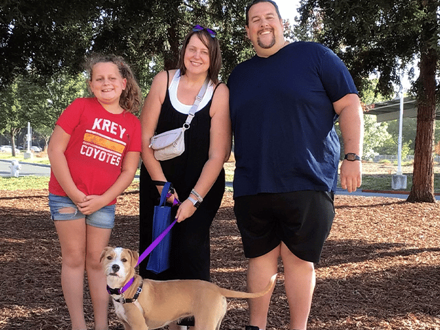 A family poses together outdoors, featuring a young girl in a red shirt, a woman in a black dress, and a man in a blue t-shirt. They are standing near a brown dog wearing a purple collar. This image highlights their recent adoption of a pet.