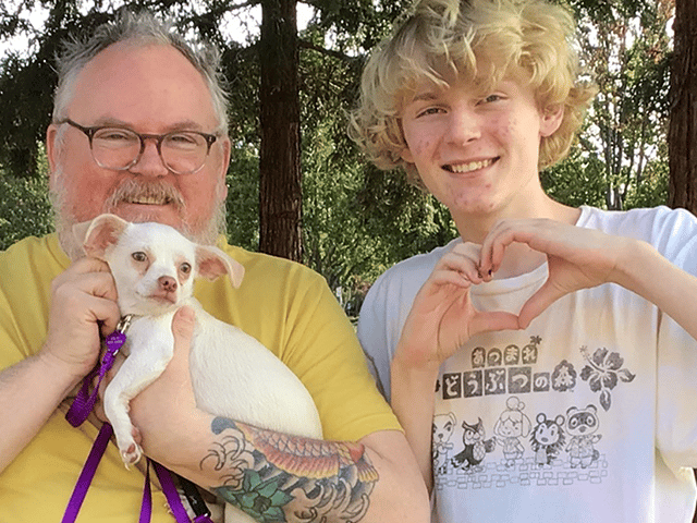 A man with glasses holding a small white dog and a teenager making a heart shape with his hands, standing outdoors among trees. The image emphasizes the bond between them, showcasing the joy of adopted pets.