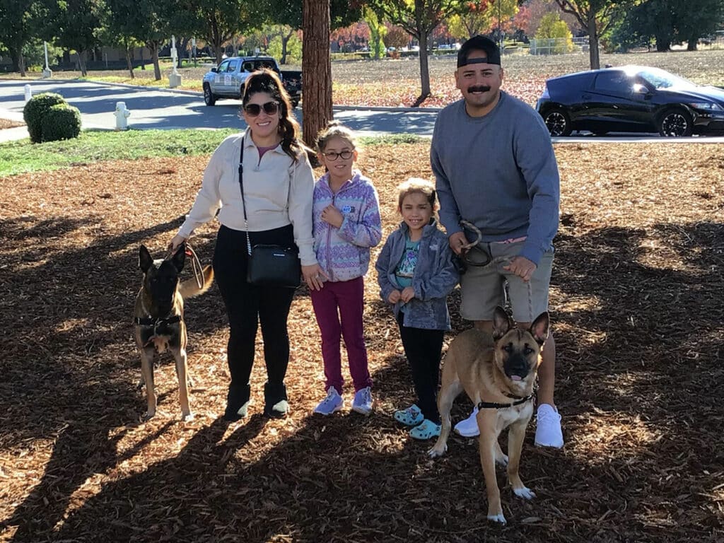 A family of four stands in a park covered with mulch, holding two leashed dogs. The adults are wearing casual attire, and two children are in colorful clothing. Sunlight filters through the trees, creating a pleasant atmosphere. This photo captures an adopted pet experience.