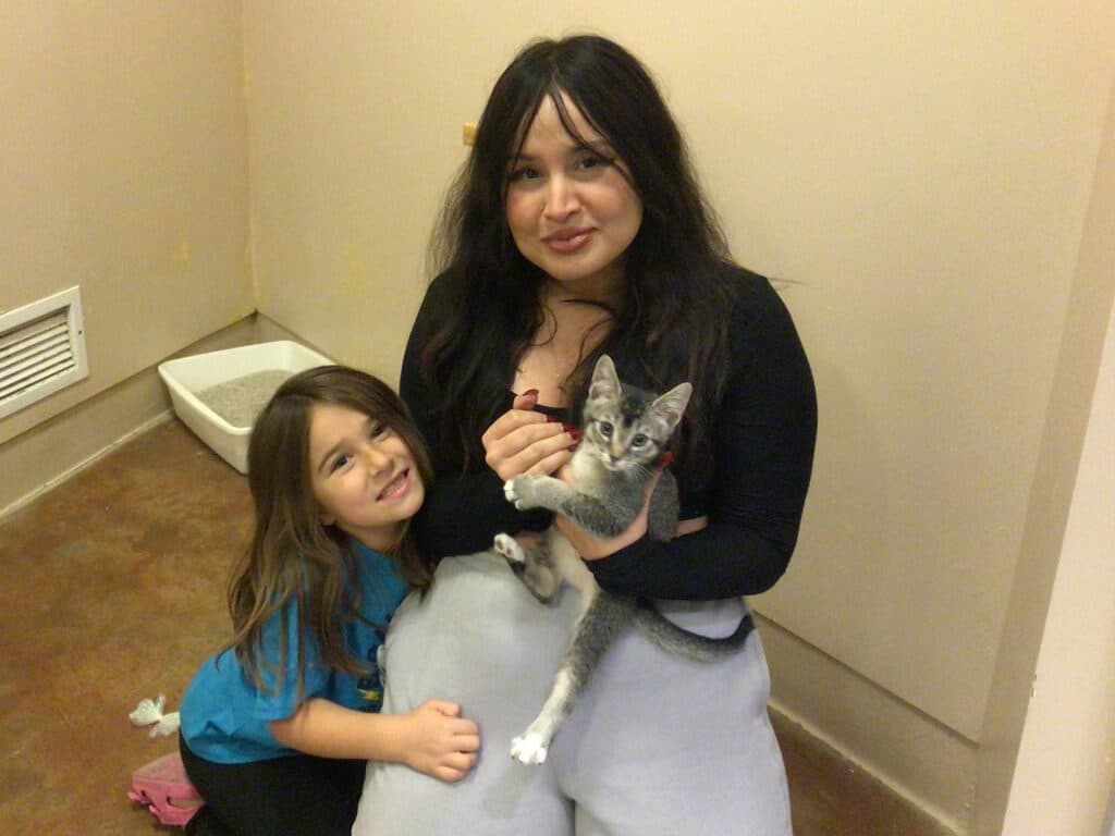 A woman with dark hair and a black top holds a gray kitten, while a young girl in a blue shirt smiles beside her, in a room with light-colored walls and a litter box in the background. This image represents an adopted pet.
