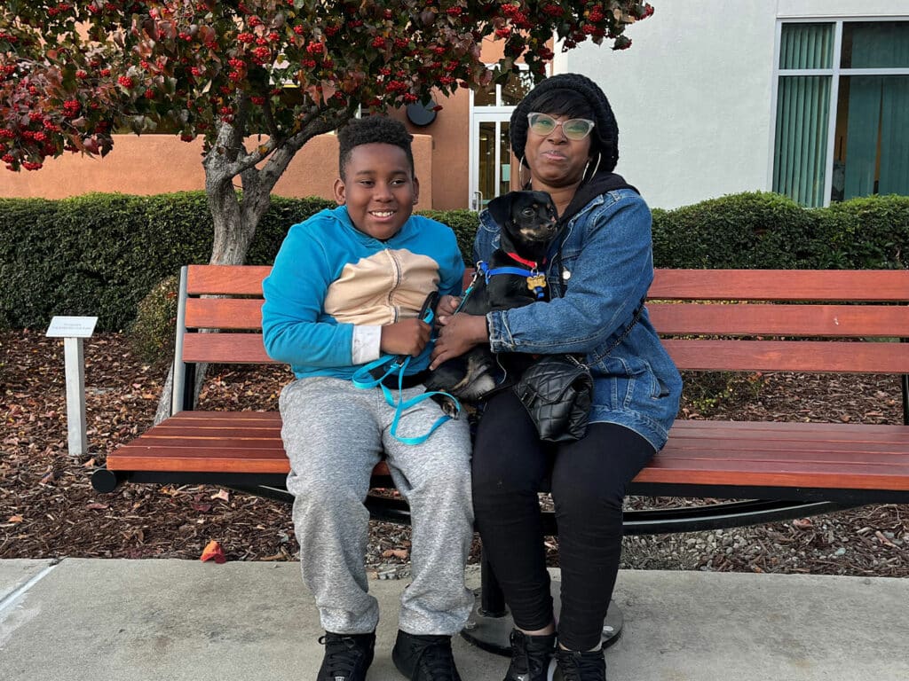 A woman and a boy sitting on a bench outside, both smiling and holding an adopted pet dog. The boy is in a blue hoodie and gray sweatpants, while the woman wears a black jacket with a hood. They are surrounded by greenery and autumn leaves.