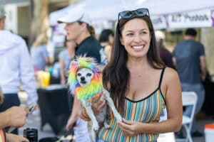 A woman smiling while holding a small dog with a colorful, rainbow wig, standing in a lively outdoor event with tents and people in the background. The setting reflects a community gathering where pets are celebrated, highlighting the theme of a Pet Walk.