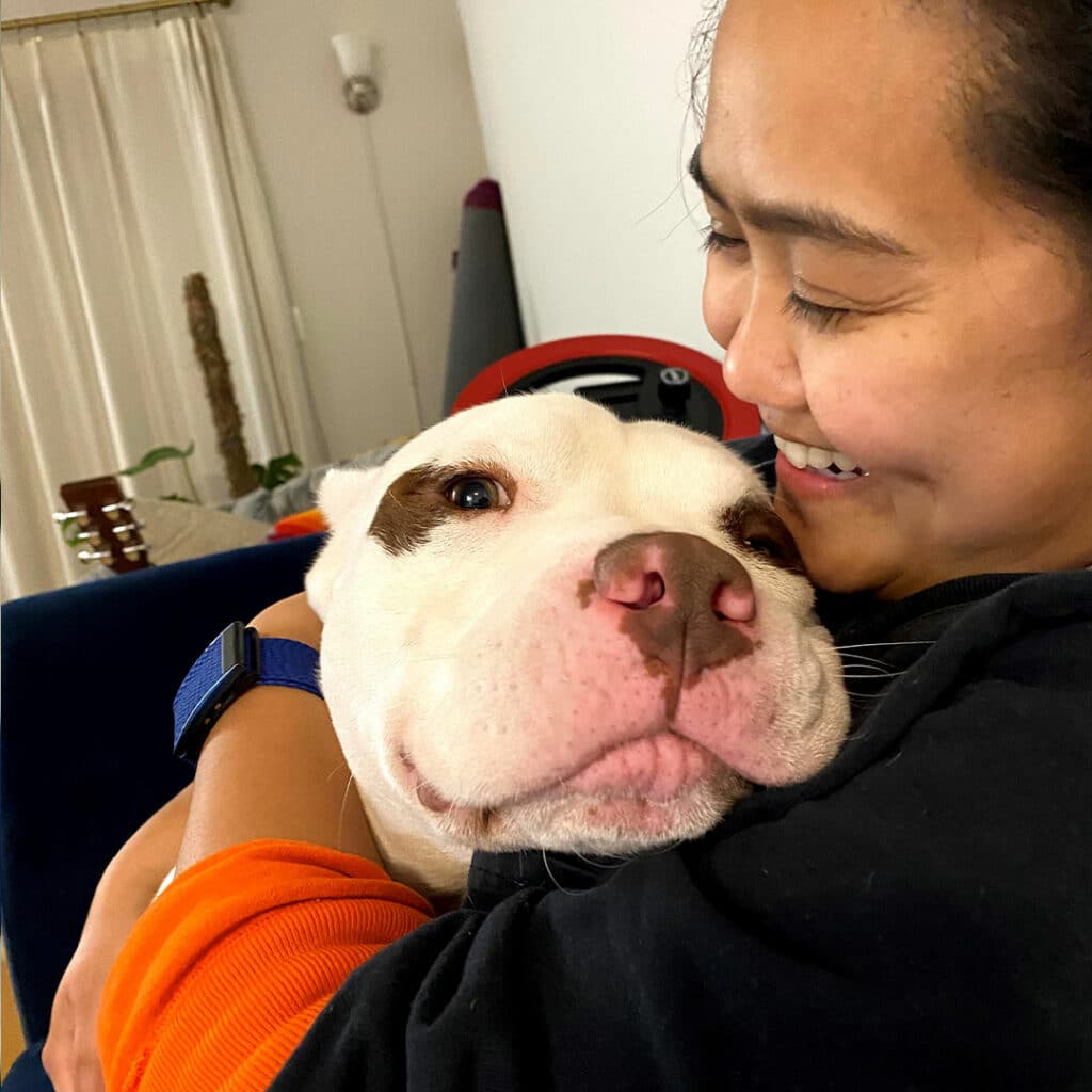 A woman smiling while holding an adopted pet dog close to her face. The dog, a white breed with brown spots, looks content in her embrace. They are in a cozy indoor setting with a guitar and plants visible in the background.