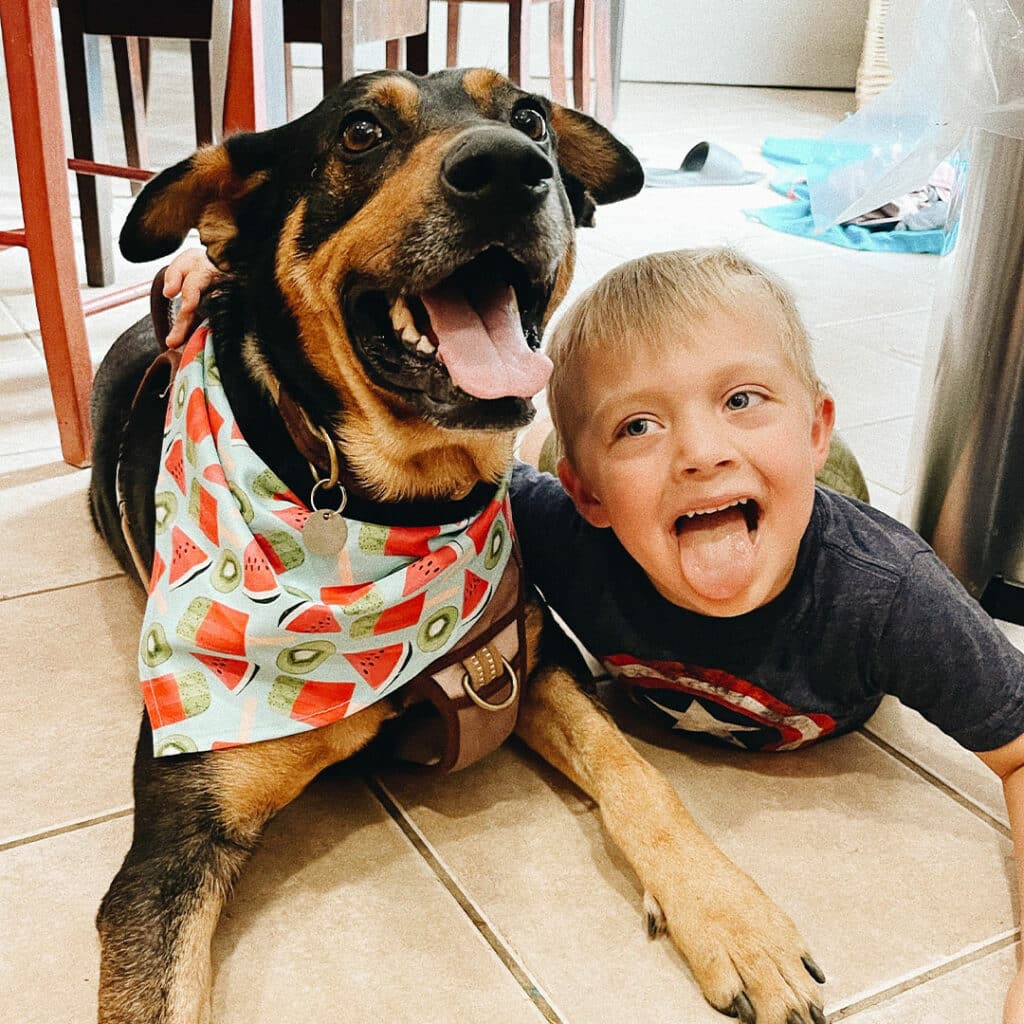 A boy lying next to a dog on tiled flooring. The dog, wearing a colorful scarf with watermelon patterns, has its tongue out, while the boy, smiling and sticking out his tongue, wears a dark t-shirt. The setting appears to be a kitchen.