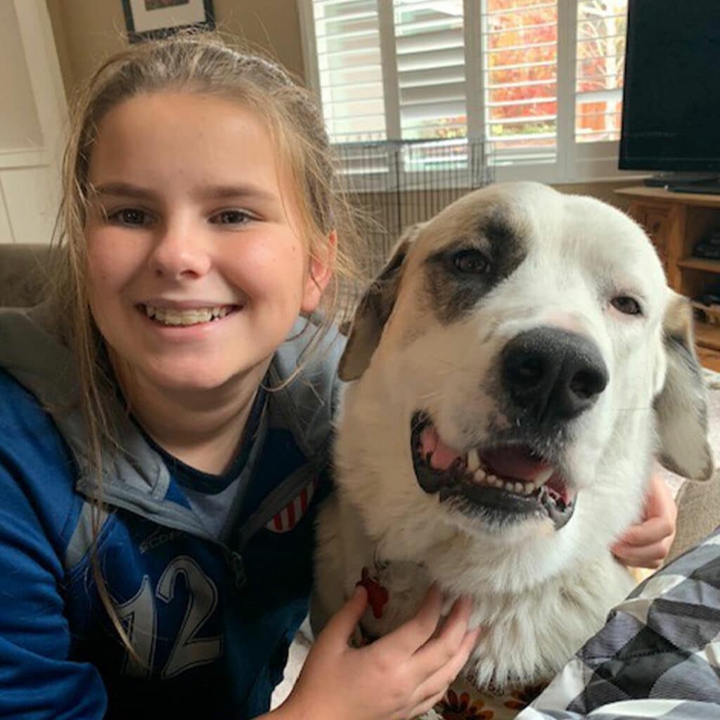 A smiling girl sits on a couch with her adopted pet dog, who has a white coat and a black patch over one eye. Both the girl and the dog are looking at the camera, creating a warm and friendly atmosphere in a living room setting.