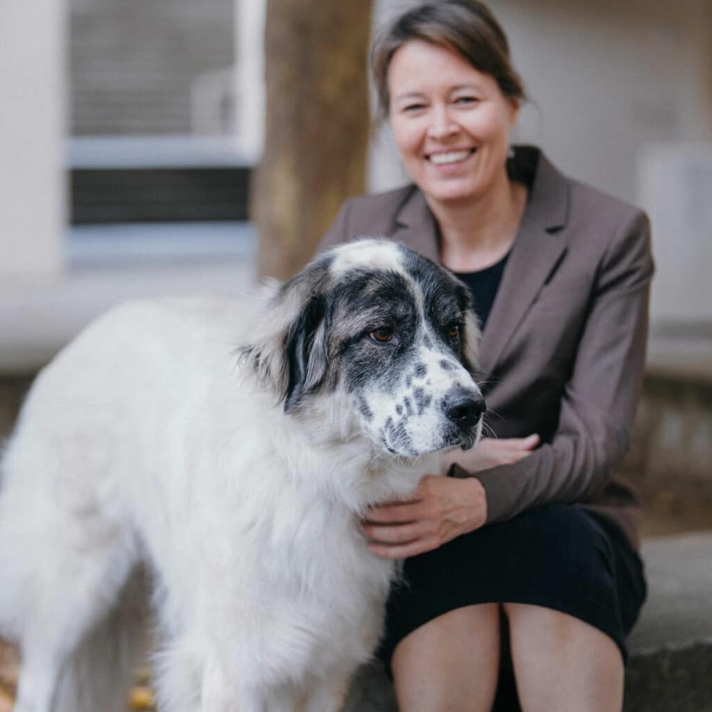A woman in a brown blazer sits on a stone step next to a large white dog with a spotted face. The woman smiles at the camera, presenting a friendly atmosphere, alongside her adopted pet.