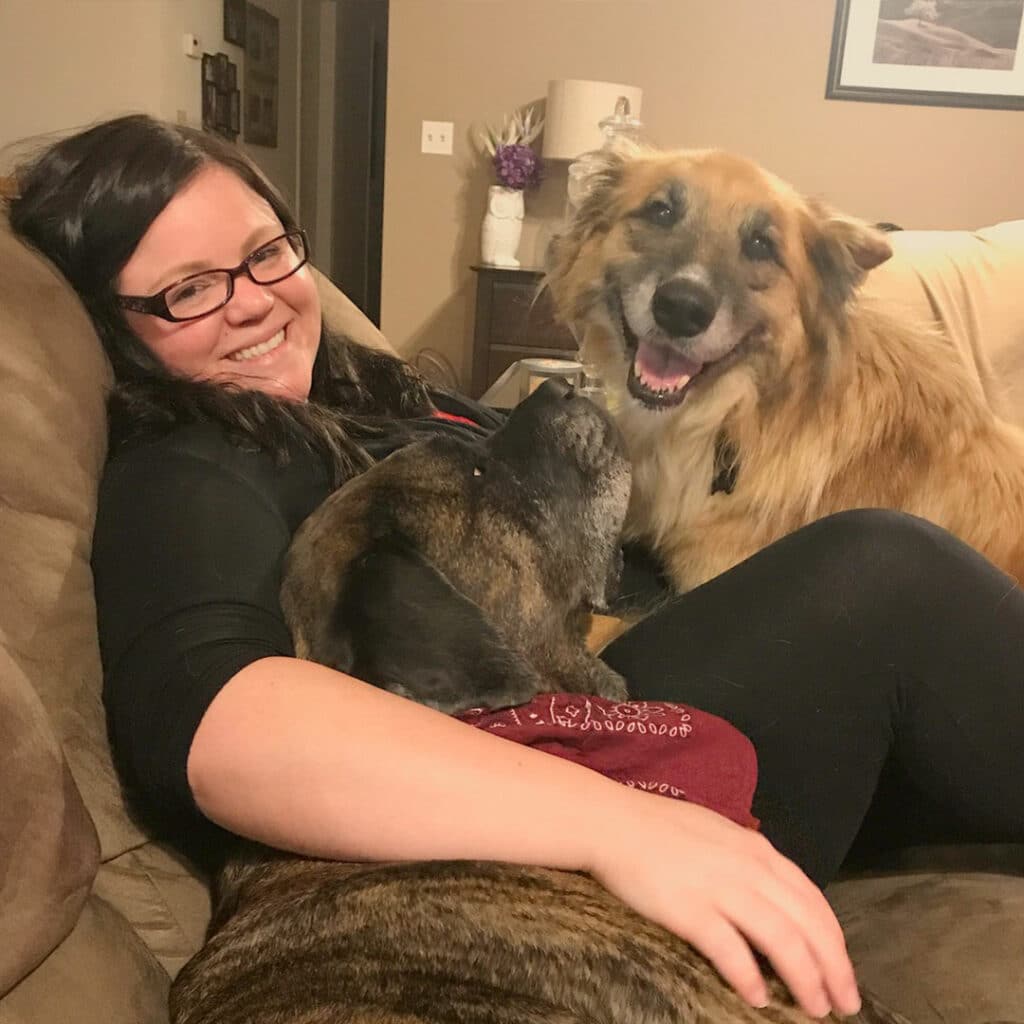 A woman with glasses sitting on a couch, smiling while two dogs are cuddled with her. One dog is a large brown and black breed, and the other is a golden-colored dog. The setting is a cozy living room.
