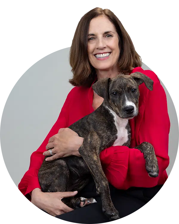 A woman with brown hair, wearing a red blouse, smiles while holding a brindle puppy in her lap. The image shows a neutral background and is framed in a circular crop, representing the Joybound Team.