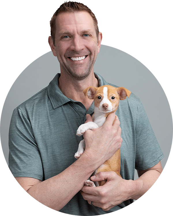 A man smiling while holding a small mixed-breed puppy. He is wearing a light green polo shirt, and the background is a soft gray, creating a neutral setting. This image supports the Joybound Team on the 'Meet the Team' page.