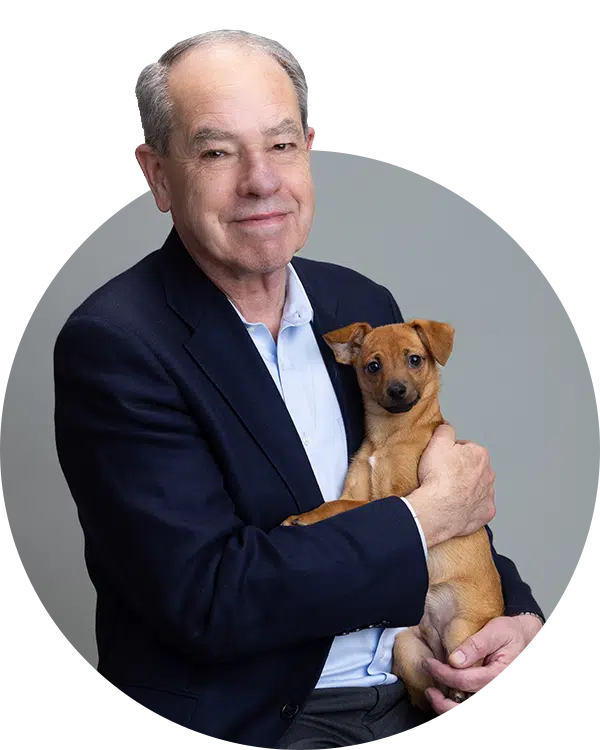 A man in a navy blazer is seated, holding a small brown dog in his arms. The background is a neutral gray color. This image showcases a member of the Joybound Team, highlighting a connection with a pet.