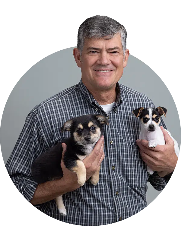 A man with gray hair wearing a plaid shirt holds two small puppies, one black and brown, the other white and brown. The background is gray, and the image is framed in a circular shape. This image represents the Joybound Team.