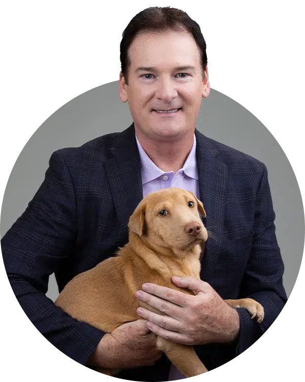 A man in a blazer holds a light brown dog while smiling at the camera. The background is a neutral gray. This image is associated with the Joybound Team, highlighting a friendly atmosphere.