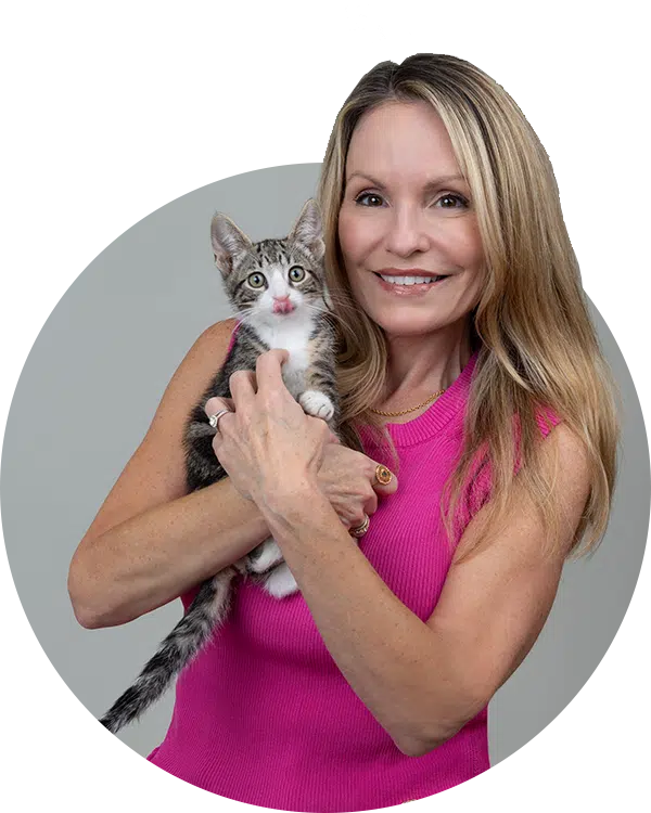A woman with long blonde hair holds a gray and white kitten in her arms. She wears a pink sleeveless top and smiles gently. The background is a soft gray, and the image is framed in a circular shape. This image showcases a member of the Joybound Team.