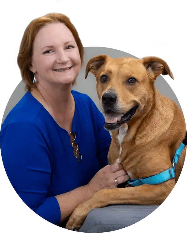A woman with medium-length hair, wearing a blue shirt, smiles while sitting beside a brown dog with a light blue harness. They appear relaxed and comfortable in a neutral background, representing the Joybound Team.
