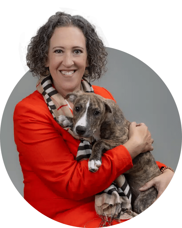 A smiling woman wearing an orange blazer and a striped scarf holds a small brown and white dog in her arms. The background is a light gray, and the image is circular, highlighting both the woman and the dog. This photo represents the Joybound Team.