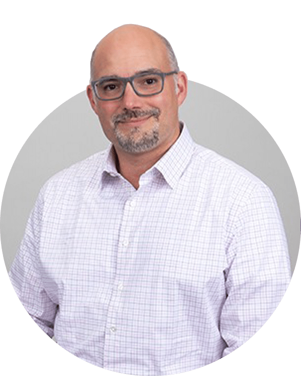 A man with glasses and a light plaid shirt stands in front of a neutral gray background. His friendly expression conveys confidence. This image represents a member of the Joybound Team, featured on the 'Meet the Team' page.