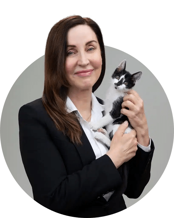 A woman in a black blazer smiles while holding a small black and white kitten. The background is neutral, and the image is framed in a circular border. This image represents the Joybound Team.