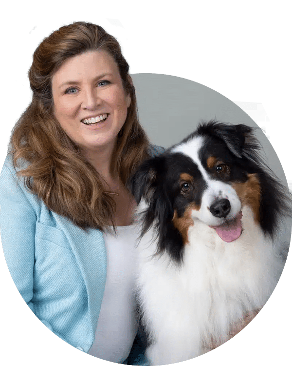 A smiling woman with long brown hair wearing a light blue jacket poses beside a fluffy dog with black and white fur. They are set against a neutral background, reflecting a friendly impression, showcasing the Joybound Team.