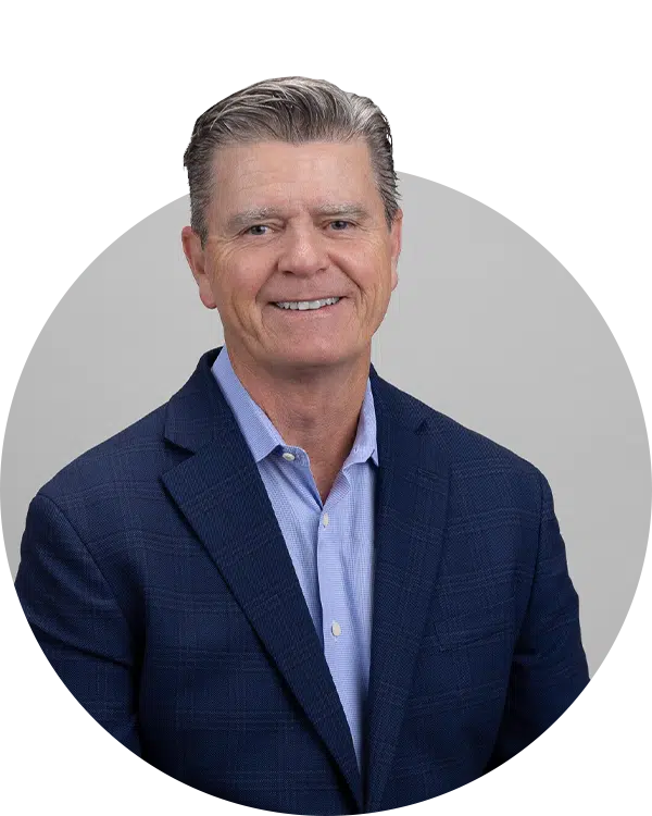 A portrait of a man with short gray hair, wearing a blue blazer and light blue shirt, smiling at the camera. The background is gray, and the image is cropped in a circular shape. This photo is part of the Joybound Team section.