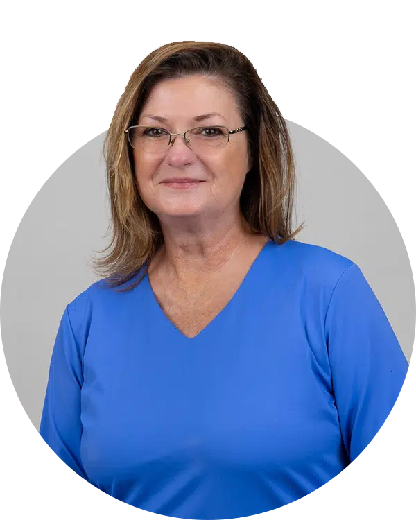 A woman with shoulder-length wavy hair, wearing glasses and a blue top, smiles in a neutral setting. This photo is part of the Joybound Team profile, showcasing team members.