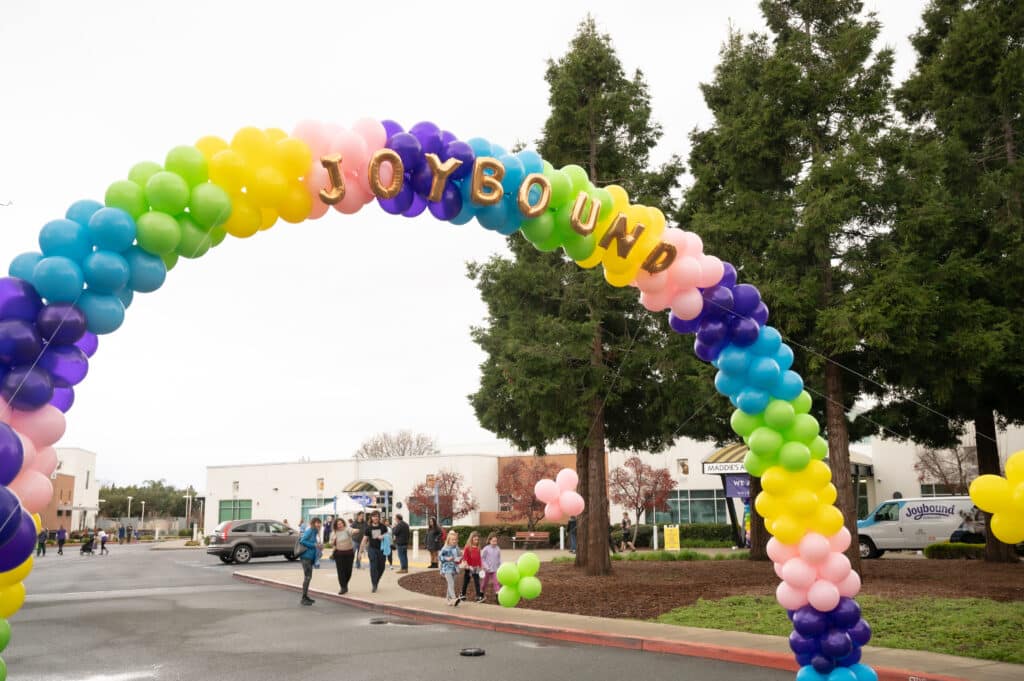 A colorful balloon arch in shades of green, pink, purple, and blue with the word "JOYBOUND" spelled out in large letters. Underneath, a few people walk on a paved path lined with trees, leading to a festival area in the background.