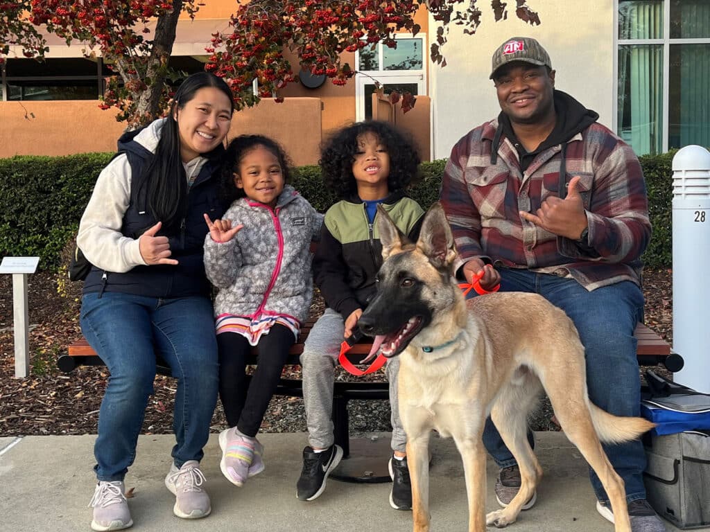 A family seated on a bench outdoors with a dog. The family includes a woman and two children, one girl and one boy, smiling and posing. The dog, a light-colored German Shepherd, sits beside them. Trees with red leaves are in the background. This image represents adoption.