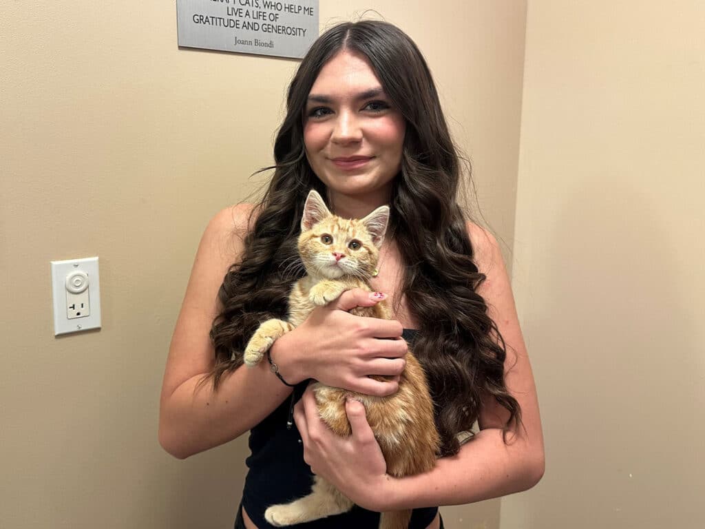A young woman with long brown hair smiles while holding a small orange kitten in her arms. The background features a wall with a quote, and there is a light switch and electrical outlet visible. This image highlights a recent adoption event.