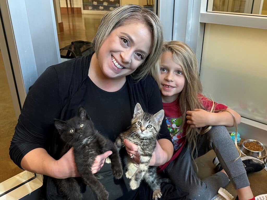 A woman and a young girl sitting on the floor in a shelter, both smiling. The woman holds a black kitten and the girl holds a gray striped kitten. Adoption appears to be the focus, with food bowls in the background.