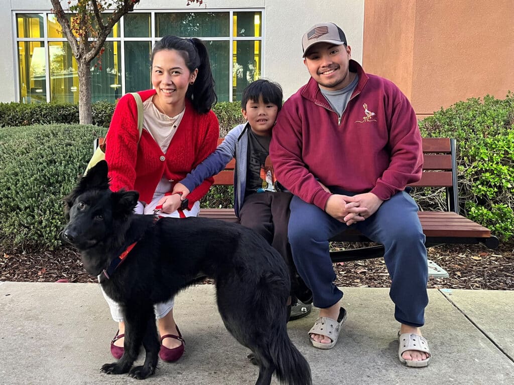 A family poses for a photo in a park. A woman wearing a red cardigan sits next to a boy and a man who is wearing a maroon top. A large black dog with a red collar sits at their feet, all smiling and relaxed, in front of a building.