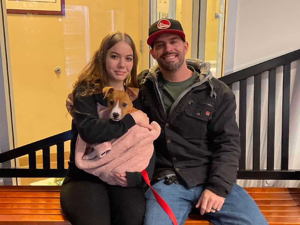A young woman and a man sitting on a bench in an indoor setting, smiling. The woman holds a small dog wrapped in a pink blanket. The backdrop shows a fenced area, suggesting an adoption event.
