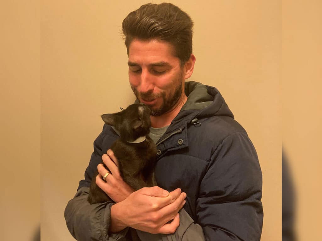 A man in a blue jacket lovingly holding a black cat close to his face. The cat appears to be affectionately nuzzling against him. The background is a simple, light color, suggesting a cozy atmosphere. This image represents recent adoption.