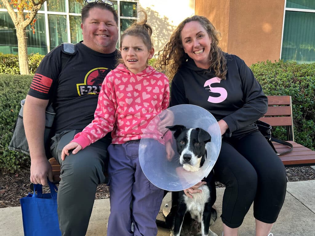A family sits on a bench outside, smiling together. A young girl in a pink heart-patterned sweater sits between two adults. One adult is wearing a black shirt, and the other wears a dark hoodie. A dog wearing an Elizabethan collar is on the girl's lap, illustrating a heartwarming moment related to adoption.