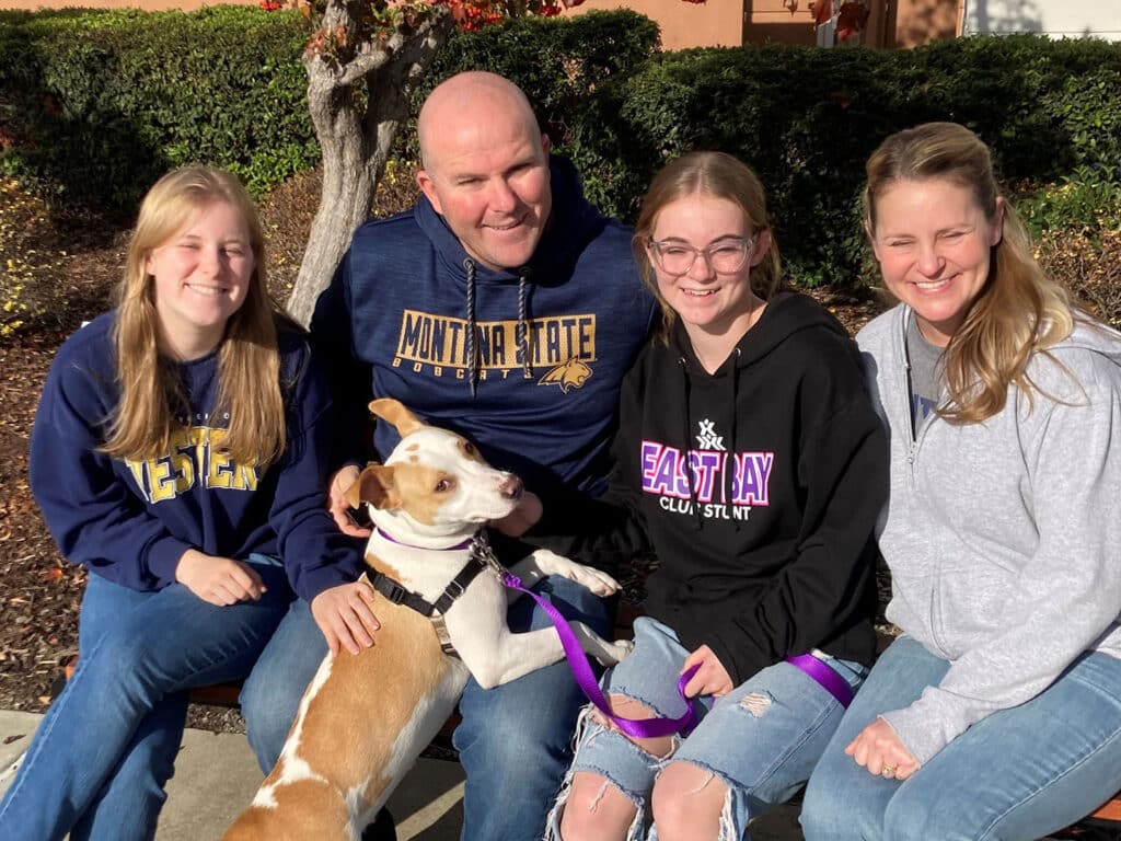 A family of four, including two teenage girls and two adults, is sitting outside. One adult holds a dog on a leash. The dog is a small, white and brown mixed breed. They are all smiling and wearing casual clothing, with trees and shrubbery in the background, highlighting a recent adoption.