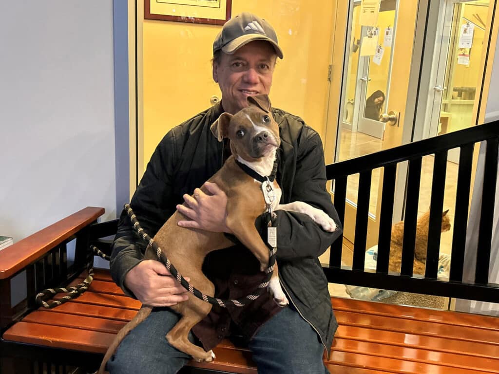 A man sitting on a wooden bench holds a small, brown dog in his arms, both looking at the camera. The setting appears to be an adoption center, with a doorway and some animal habitats visible in the background. The image highlights the theme of adoption.