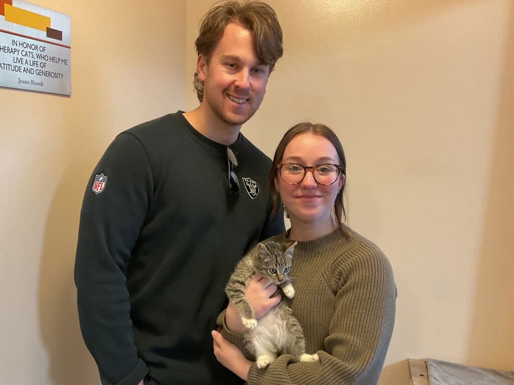 A smiling couple stands together in a room holding an adopted gray kitten. The man wears a black sweatshirt, and the woman has glasses and a green sweater. The background features a wall with a plaque.