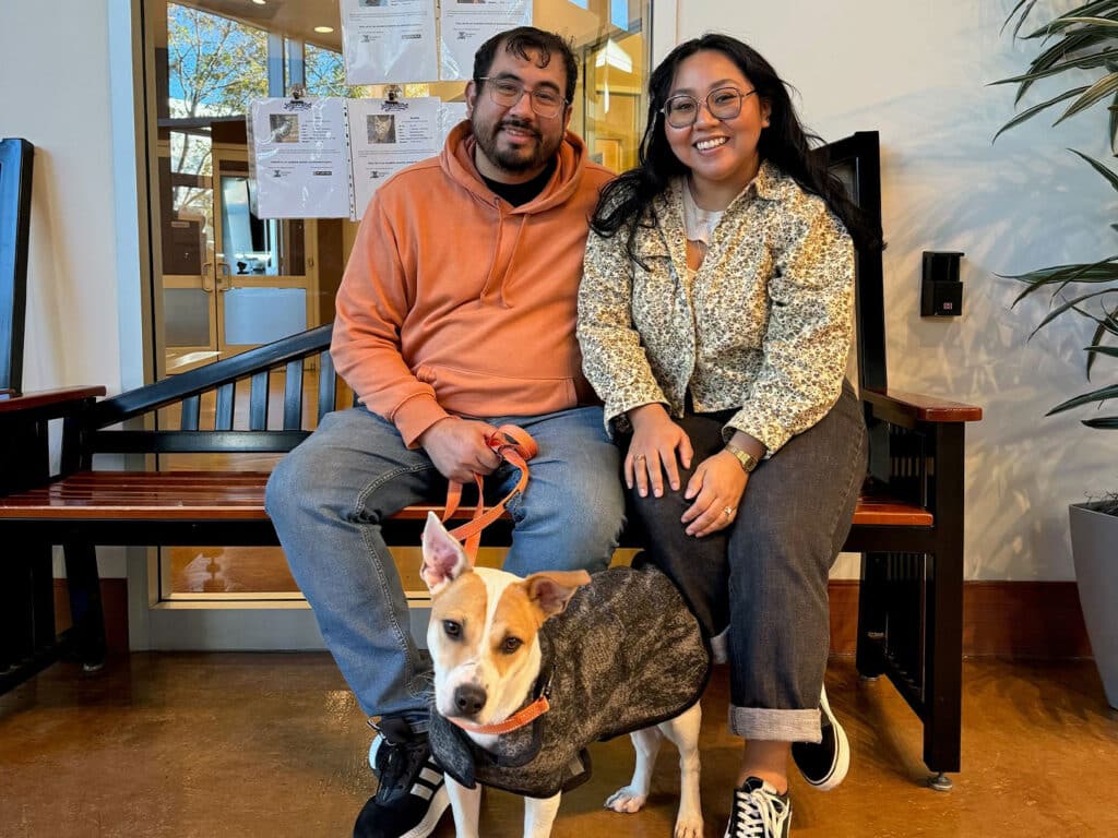 A man and a woman sit on a bench inside an adoption center with a dog between them. The woman wears glasses and a patterned jacket, while the man wears an orange hoodie. The dog, wearing a gray coat, appears relaxed, highlighting a recent adoption.