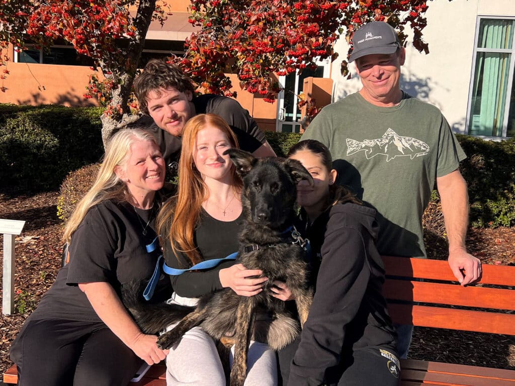 A family of five, including three adults and two children, is gathered around a medium-sized dog on a park bench. The dog, a black and tan mixed breed, is being held by a young girl. The group is smiling in a setting with colorful autumn leaves.