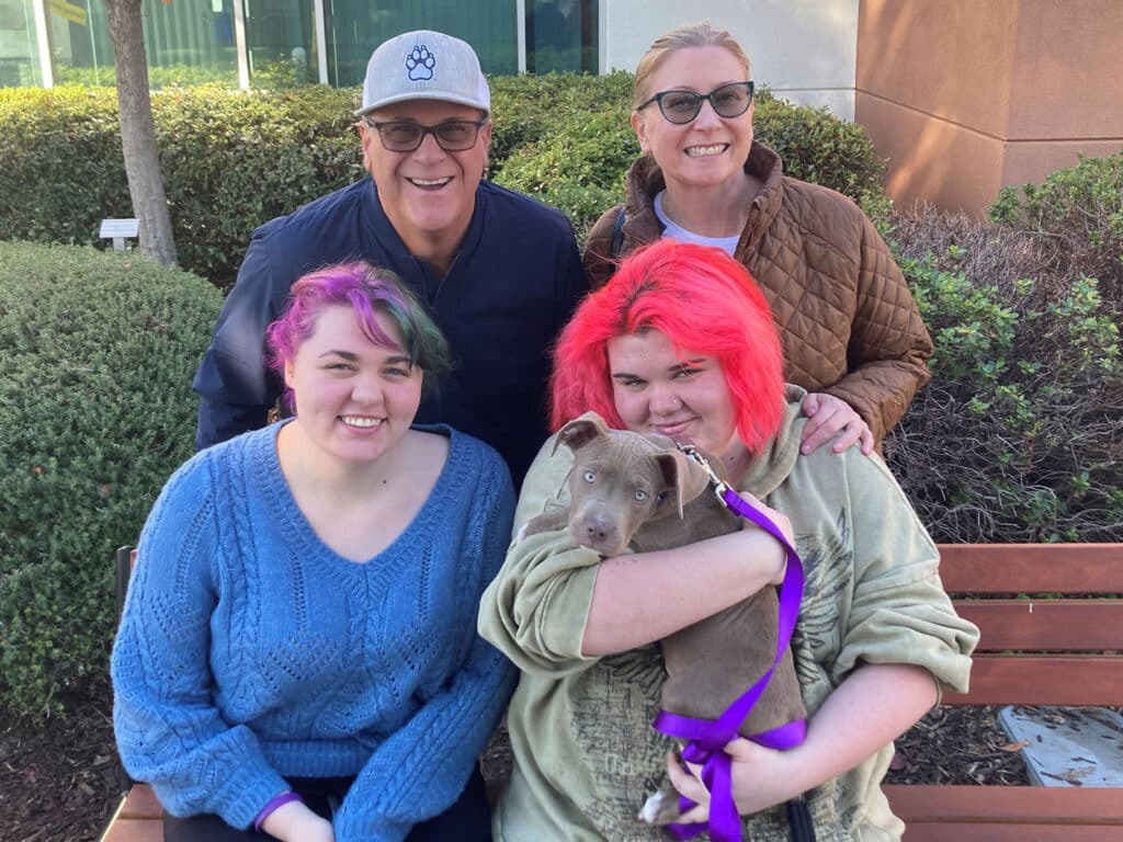 A family of four smiles together outdoors, sitting on a bench. Two young women, one with colorful hair, hold a small gray dog. Two adults stand behind them, showcasing a joyful moment of adoption.