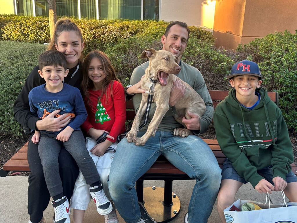 A family of five sits on a park bench, smiling and posing with a recently adopted dog. The children, dressed in casual attire, are seated close together, while the parents hold the dog, highlighting the joy of adoption.