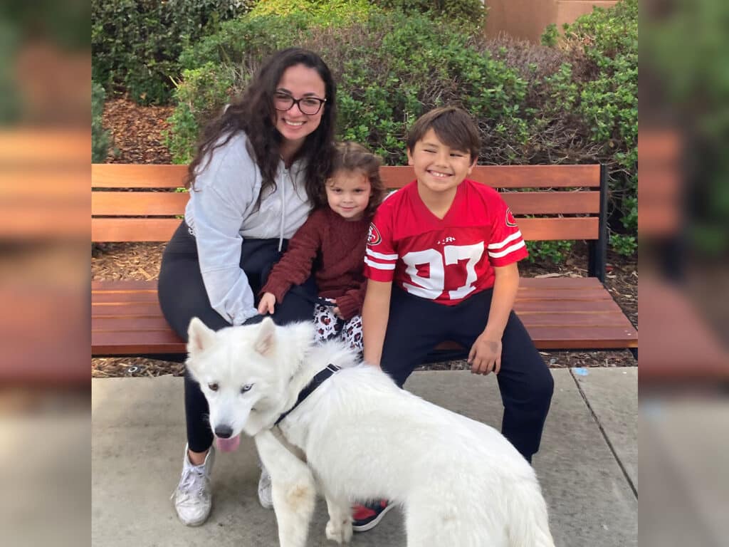 A woman with long dark hair sits on a bench beside two children, a girl and a boy. The girl is in red and gray, while the boy wears a red jersey with the number 97. A white dog stands in front of them, contributing to a joyful scene related to adoption.