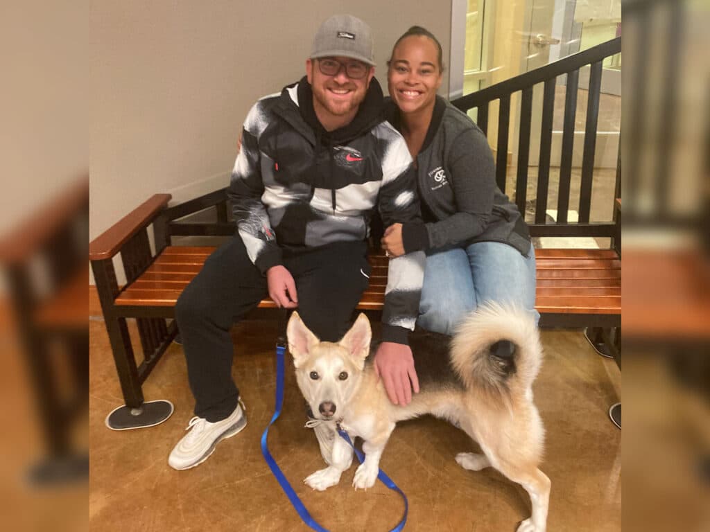 A man and woman sitting together on a wooden bench in a welcoming indoor space, smiling with a dog beside them. The dog, with a light coat and a bushy tail, is leashed and looking towards the camera. This image highlights a recent dog adoption.