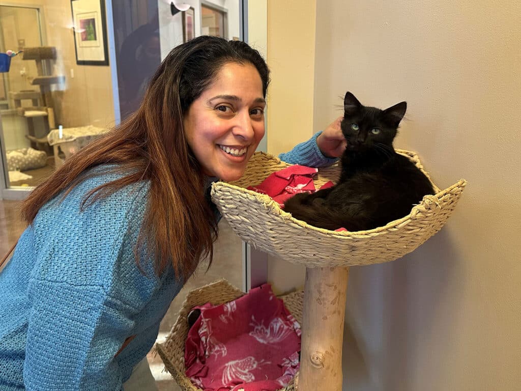 A woman with long hair smiles while leaning next to a black cat resting in a woven cat bed. The setting appears to be an animal adoption center with a colorful backdrop and visible pet habitats.