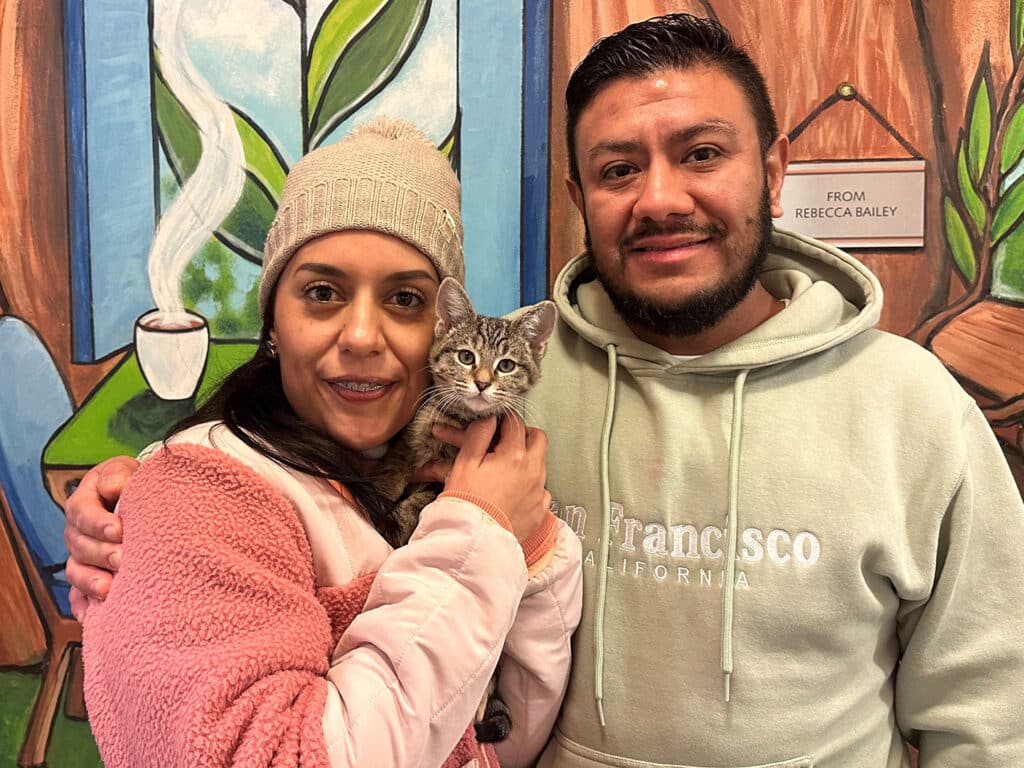 A couple stands in front of a colorful mural holding a small kitten. The woman, wearing a pink jacket and a beige beanie, smiles as she cradles the kitten. The man beside her wears a light hoodie. This image highlights their recent adoption.