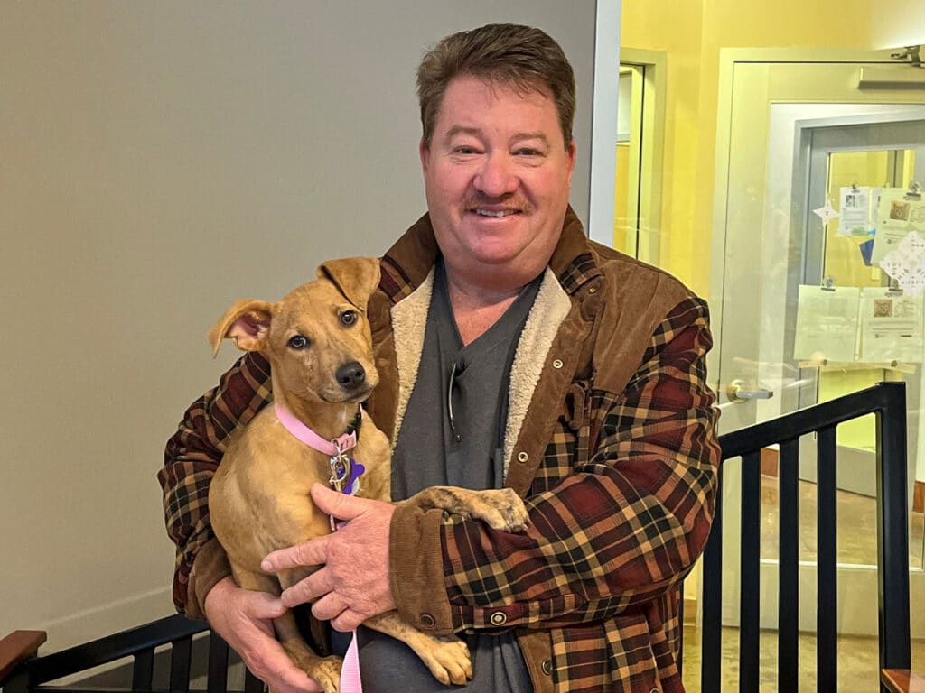 A man holding a tan puppy with floppy ears in a cozy indoor setting. The man is wearing a plaid jacket and smiling, while the puppy has a pink collar. This image represents an adoption event from the week of December 9.