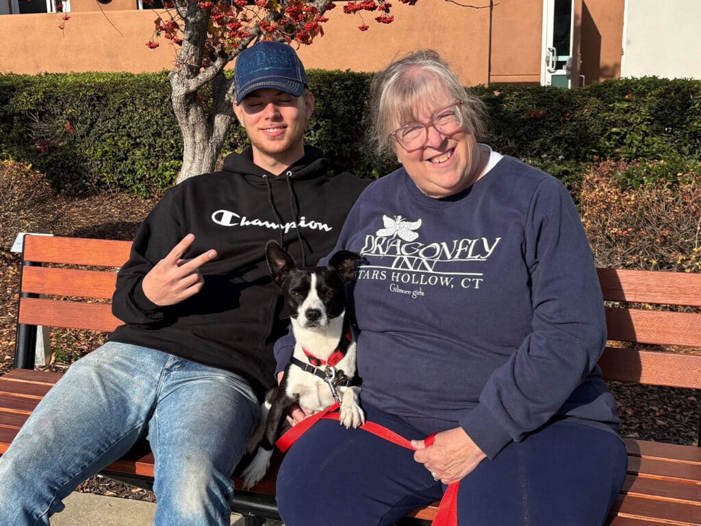 A young man and an older woman smiling while seated on a bench outdoors, with a small dog in front of them. The woman wears a blue sweatshirt, and the man is in a black hoodie. The setting includes shrubbery and a building in the background. Adoption is highlighted through their interaction.
